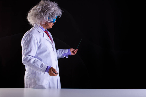 Szalony nauczyciel fizyki w białym płaszczu z rozczochranymi włosami w śmiesznych okularach, trzymający różdżkę, by wskazać na tablicę.  - Zdjęcie, obraz