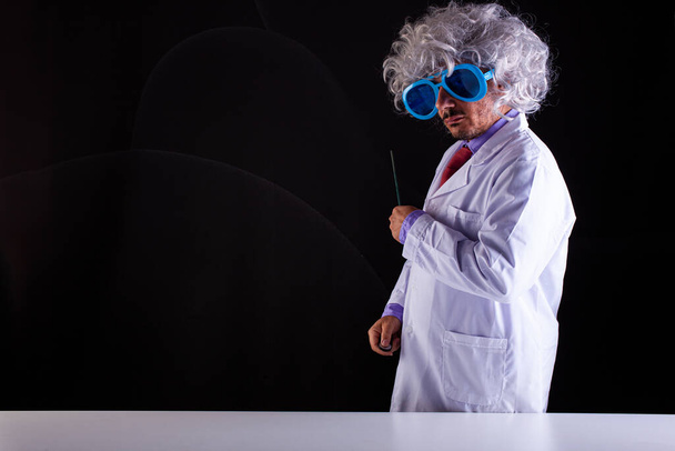 Crazy science teacher in white coat with unkempt hair in funny eye glasses holding a wand to point at the blackboard  - 写真・画像