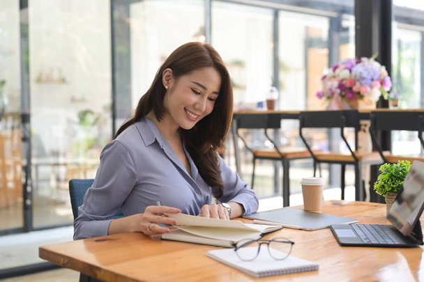 Smiling businesswoman working with computer tablet and making note on notebook. - 写真・画像