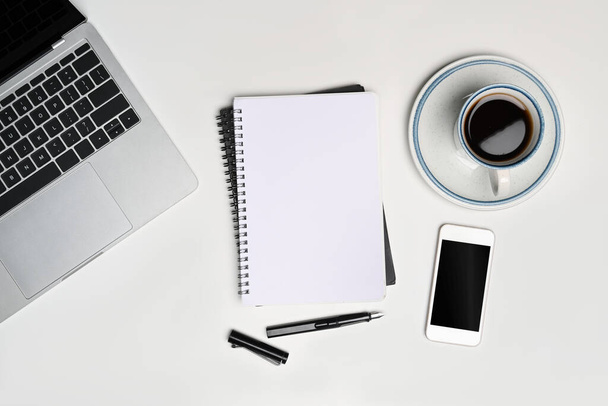 Empty notebook, mobile phone, coffee cup and laptop on white desk. - Photo, Image