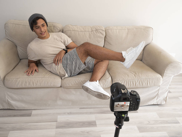 Hispanic young man creating clothing content for social networks with a camera on a tripod, sitting on a piece of furniture - Photo, image