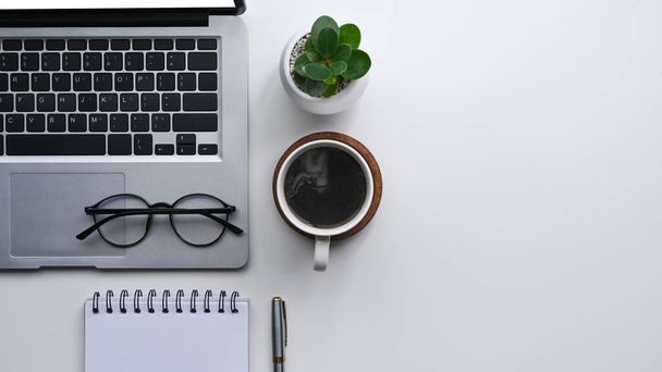 Einfacher Arbeitsplatz mit Computer-Laptop, Kaffeetasse, Brille, Notizbuch und Pflanze auf weißem Schreibtisch. - Foto, Bild