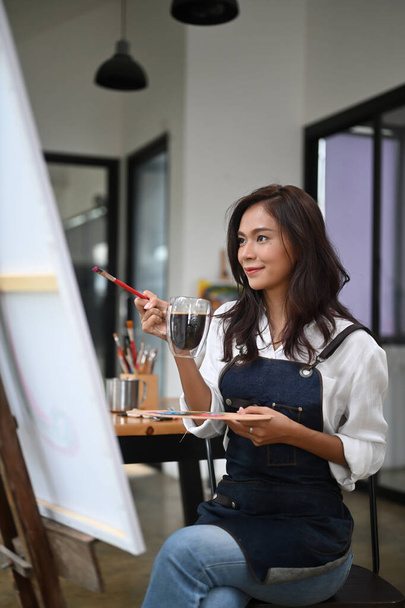 Beautiful creative woman drinking coffee while sitting at her art workshop. - Фото, зображення