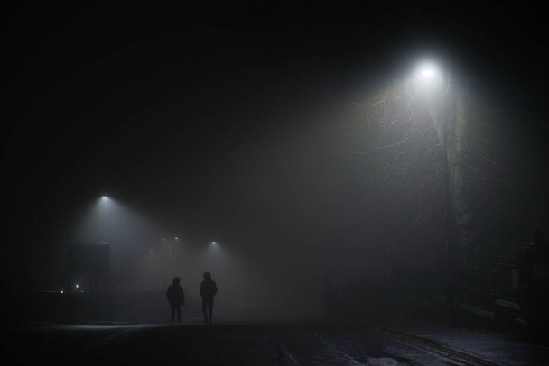 Міська зимова сцена двох чоловіків, що ходять під вуличними вогнями в надзвичайно туманних умовах. Мужність або жахливе відчуття, обидва носять чорні пальто з капюшоном вгору
. - Фото, зображення