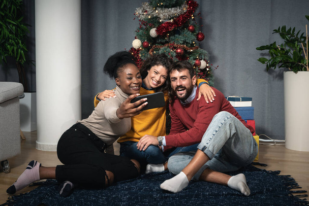 Drei junge Freunde feiern gemeinsam Weihnachten und Silvester, indem sie Witze machen und Selfies mit dem Smartphone vor dem Weihnachtsbaum machen und bis Mitternacht darauf warten, die Geschenke in Schachteln zu tauschen. - Foto, Bild