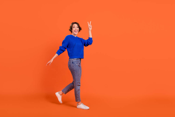 Full length body size view of attractive cheerful girl walking showing v-sign isolated over bright orange color background - Foto, immagini