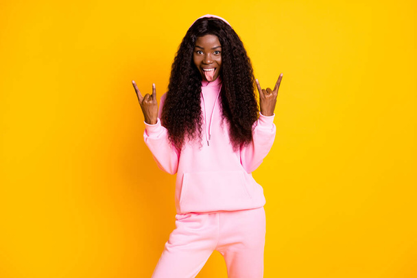 Photo of shiny cool dark skin woman dressed pink hoodie showing tongue finger hand rock sign isolated yellow color background - Zdjęcie, obraz