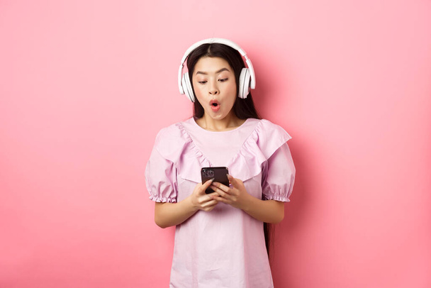 Aufgeregtes asiatisches Mädchen schaut amüsiert auf Smartphone-Bildschirm, sagt wow, hört Musik in drahtlosen Kopfhörern, steht vor rosa Hintergrund - Foto, Bild