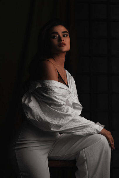 Piękna młoda dziewczyna w białych ubraniach pozowanie w ciemnym studio fotograficznym. Zdjęcie poniżej pasa. Wrażliwa brązowłosa kobieta z długimi włosami - Zdjęcie, obraz
