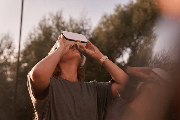 A menina, usando óculos de realidade virtual, segura os óculos com as mãos e olha para o topo. Contra o fundo de árvores verdes. Há um feixe de luz solar nas proximidades. - Foto, Imagem