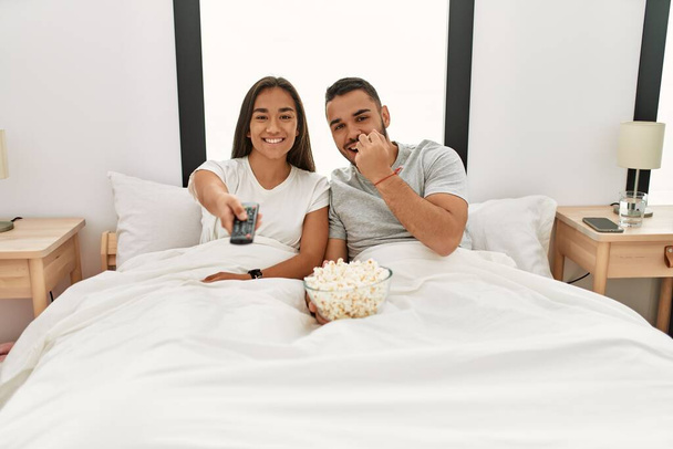 Junges lateinisches Paar schaut Film und isst Popcorn liegend auf dem Bett. - Foto, Bild