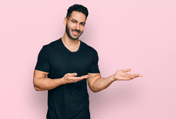 Spanisch Mann mit Bart trägt lässig schwarzes T-Shirt erstaunt und lächelt in die Kamera, während er mit der Hand präsentiert und mit dem Finger zeigt.  - Foto, Bild