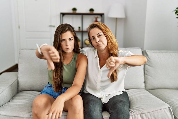 Мать и дочь вместе сидят дома на диване и выглядят несчастными и сердитыми, показывая отказ и негатив с опущенными пальцами. плохое выражение.  - Фото, изображение