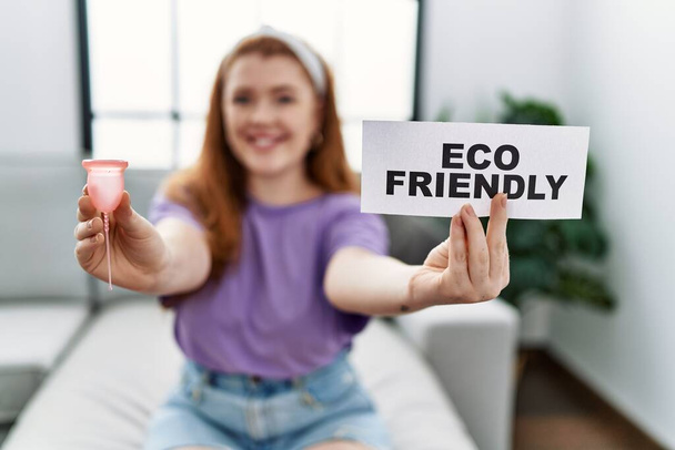 Young redhead woman smiling confident holding menstrual cup and eco friendly banner at home - Foto, Imagem