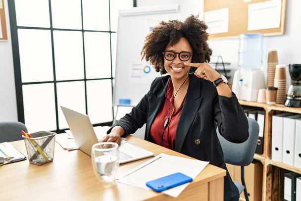Mujer afroamericana con cabello afro trabajando en la oficina con auriculares de operador apuntando con el dedo a la mano y la nariz, sonriendo alegre. concepto de belleza  - Foto, Imagen