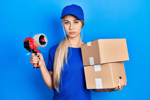 Young caucasian woman holding packages and packing tape relaxed with serious expression on face. simple and natural looking at the camera.  - Photo, Image