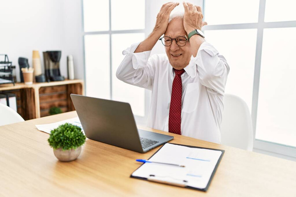 Älterer Mann, der im Büro mit Computer-Laptop arbeitet und unter Kopfschmerzen leidet, verzweifelt und gestresst wegen Schmerzen und Migräne. Hände auf den Kopf.  - Foto, Bild
