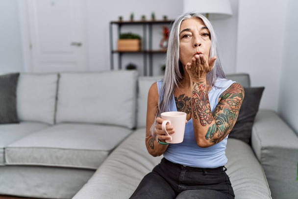 Femme aux cheveux gris du moyen âge buvant du café assis sur le canapé à la maison en regardant la caméra souffler un baiser avec la main sur l'air étant belle et sexy. expression d'amour.  - Photo, image