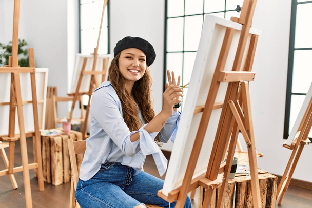 Młoda latynoska artystka malująca na płótnie w pracowni sztuki uśmiechając się pozytywnie robiąc znak ok ręką i palcami. udana ekspresja.  - Zdjęcie, obraz