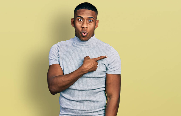 Fiatal fekete férfi visel alkalmi póló meglepett mutató ujjal oldalra, nyitott szájú csodálkozó kifejezés.  - Fotó, kép