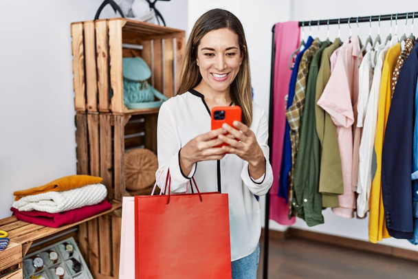 Young hispanic woman using smartphone shopping at clothing store - Zdjęcie, obraz