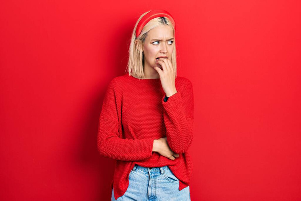 Mooie blonde vrouw draagt casual rode trui kijken gestrest en nerveus met handen op de mond bijtende nagels. angstprobleem.  - Foto, afbeelding