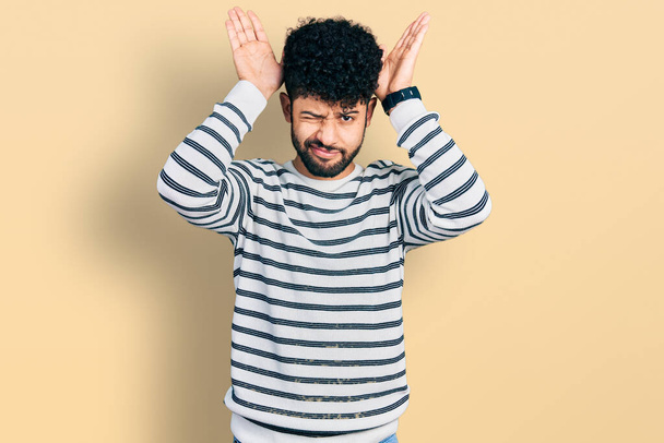 Młody arabski mężczyzna z brodą, noszący luźny sweter w paski, wykonujący gesty króliczych uszu dłońmi, wyglądającymi cynicznie i sceptycznie. koncepcja królików wielkanocnych.  - Zdjęcie, obraz