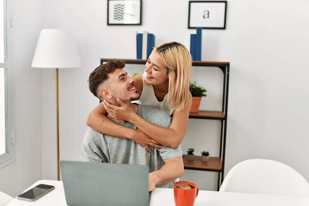 Junges kaukasisches Paar umarmt und benutzt Laptop auf dem Schreibtisch zu Hause. - Foto, Bild