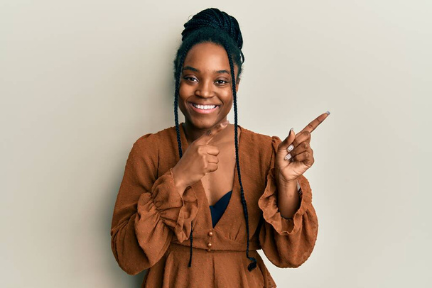 Afrikkalainen amerikkalainen nainen, jolla on punotut hiukset yllään rento ruskea paita hymyilee ja katsoo kameraa osoittaen kahdella kädellä ja sormella sivulle.  - Valokuva, kuva