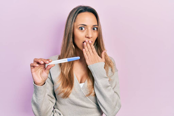 Beautiful hispanic woman holding pregnancy test result covering mouth with hand, shocked and afraid for mistake. surprised expression  - Photo, Image
