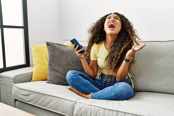 Beautiful hispanic woman sitting on the sofa at home using smartphone crazy and mad shouting and yelling with aggressive expression and arms raised. frustration concept.  - Zdjęcie, obraz