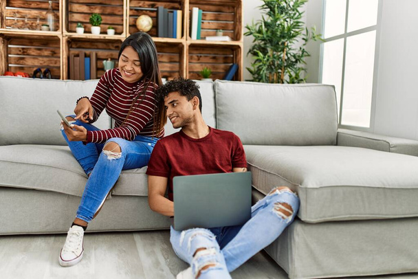 Νεαρό latin ζευγάρι χαμογελά χαρούμενος χρησιμοποιώντας φορητό υπολογιστή και smartphone στο σπίτι. - Φωτογραφία, εικόνα