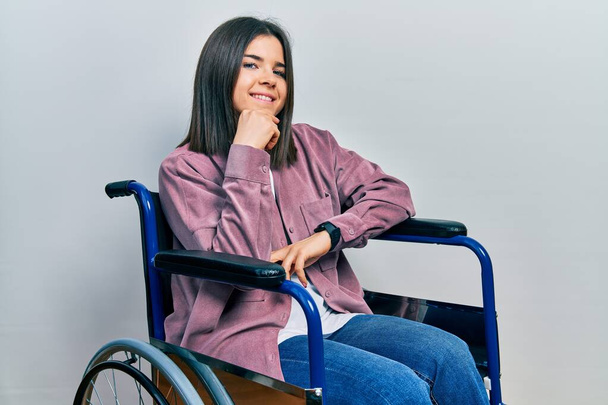 Young brunette woman sitting on wheelchair looking confident at the camera smiling with crossed arms and hand raised on chin. thinking positive.  - Foto, afbeelding