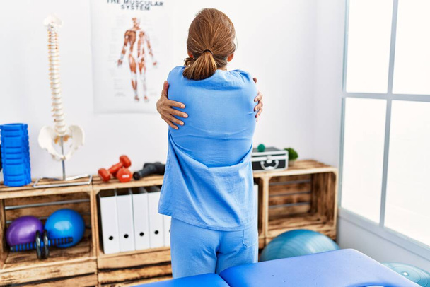 Middle age hispanic physiotherapist woman working at pain recovery clinic hugging oneself happy and positive from backwards. self love and self care  - Photo, Image