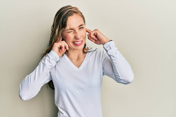 Young blonde woman wearing casual clothes covering ears with fingers with annoyed expression for the noise of loud music. deaf concept.  - Photo, Image