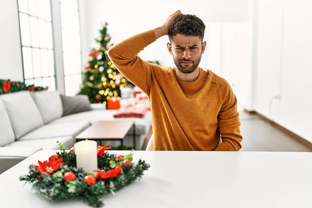 Arab young man sitting on the table by christmas tree confuse and wonder about question. uncertain with doubt, thinking with hand on head. pensive concept.  - Photo, image