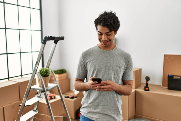 Jeune homme hispanique souriant heureux en utilisant un smartphone à la nouvelle maison. - Photo, image