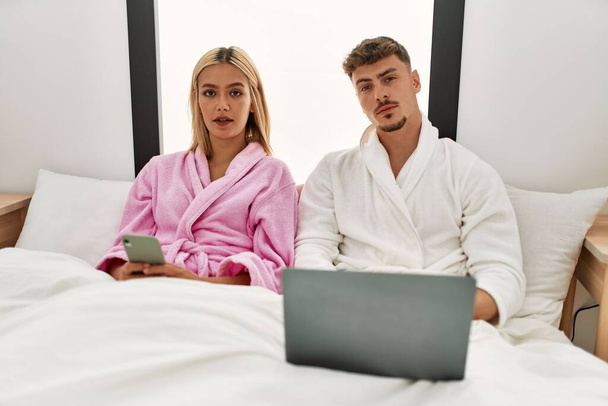Fiatal kaukázusi pár komoly kifejezés segítségével laptop és okostelefon ül az ágyon otthon. - Fotó, kép