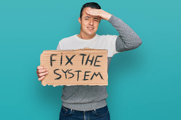 Handsome young man holding fix the system banner cardboard stressed and frustrated with hand on head, surprised and angry face  - Фото, изображение