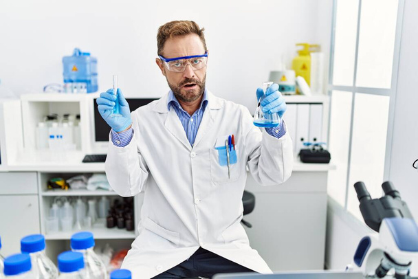 Mann mittleren Alters, der im wissenschaftlichen Labor arbeitet und chemische Produkte im Schockgesicht hält, sieht skeptisch und sarkastisch aus, überrascht mit offenem Mund  - Foto, Bild