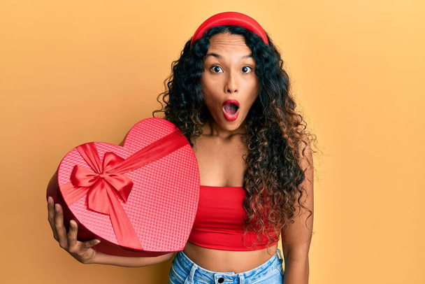 Mujer latina joven sosteniendo regalo de San Valentín asustada y sorprendida con la boca abierta para sorpresa, cara de incredulidad  - Foto, Imagen
