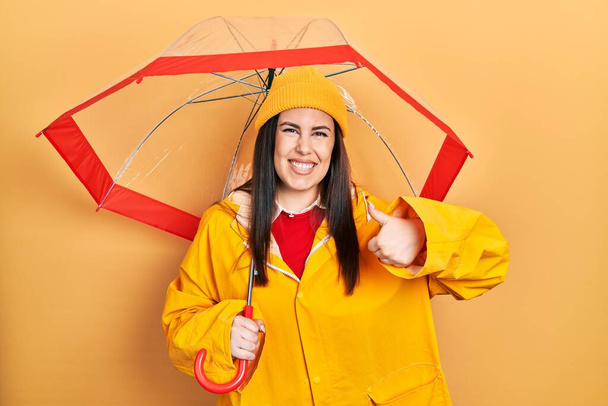 Giovane donna ispanica indossa impermeabile giallo con ombrello sorridente felice e positivo, pollice in su facendo segno eccellente e approvazione  - Foto, immagini