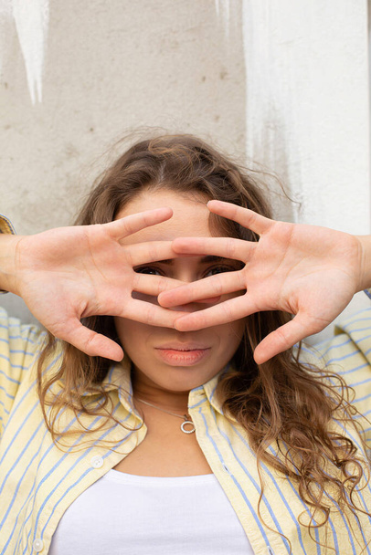 jong wit meisje met donker haar in de buurt van de muur bedekt haar gezicht met handen - Foto, afbeelding