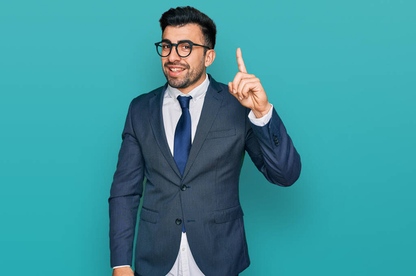 Hombre hispano con barba usando traje de negocios y corbata señalando con el dedo hacia arriba con una idea exitosa. exitado y feliz. número uno.  - Foto, imagen