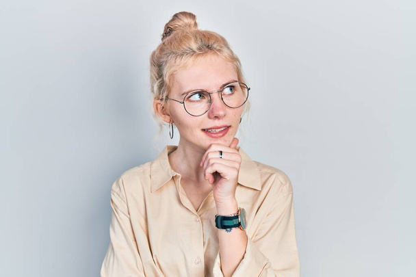 Beautiful caucasian woman with blond hair wearing casual look and glasses with hand on chin thinking about question, pensive expression. smiling with thoughtful face. doubt concept.  - Photo, Image