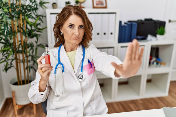 Μεσήλικας γιατρός γυναίκα κρατώντας ηλεκτρονικό τσιγάρο στην ιατρική κλινική με ανοιχτό χέρι κάνει στοπ με σοβαρή και σίγουρη έκφραση, χειρονομία άμυνας  - Φωτογραφία, εικόνα