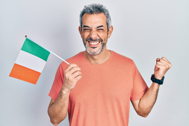 アイルランドの旗を保持している灰色の髪を持つハンサムな中年の男は誇りに思って叫んで、勝利と成功を祝う非常に興奮して腕を上げ  - 写真・画像