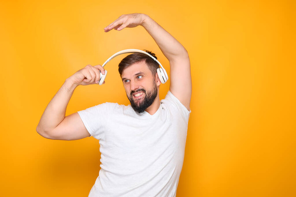 a bearded man in a white T-shirt, who stands on a yellow background, listens to music on headphones. - Photo, Image