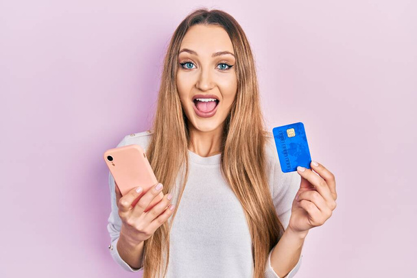 Joven chica rubia sosteniendo teléfono inteligente y tarjeta de crédito celebrando loco y sorprendido por el éxito con los ojos abiertos gritando emocionado.  - Foto, Imagen