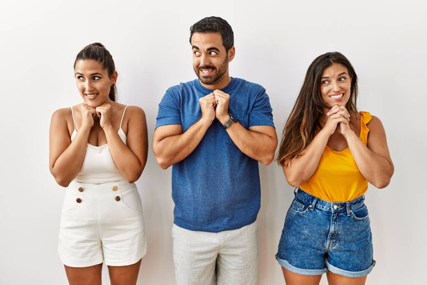 Grupo de jóvenes hispanos de pie sobre un fondo aislado riendo nerviosos y emocionados con las manos en la barbilla mirando hacia un lado  - Foto, Imagen